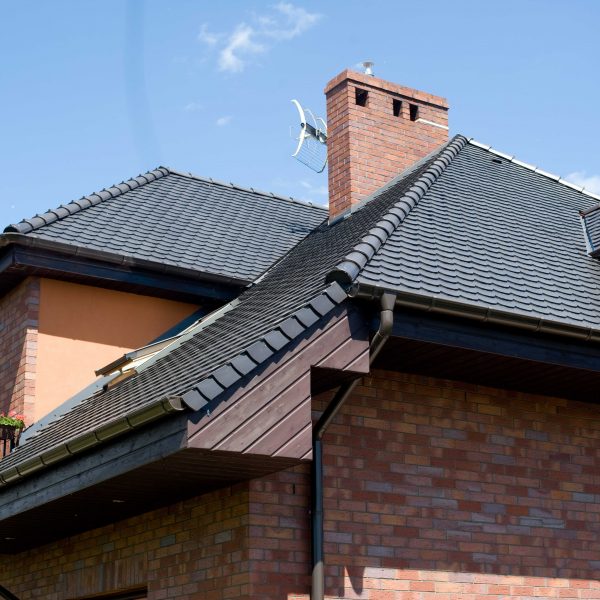 Zbliżenie na dach pokryty dachówką ceramiczną Koramic karpiówka w kolorze antracytowym. Budynek z elewacją z czerwonej cegły klinkierowej, czarnymi rynnami i elementami wykończeniowymi w ciemnym drewnie.