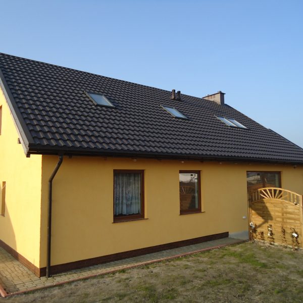 Dom z żółtą elewacją oraz dachem pokrytym blachodachówką Ruukki Finnera w kolorze brązowym, wyposażony w kilka okien dachowych oraz elementy małej architektury ogrodowej.