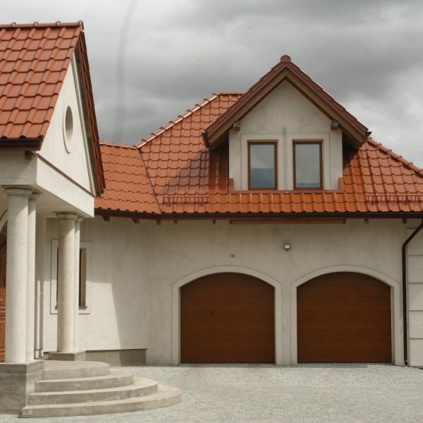 Dach domu pokryty dachówką ceramiczną Wienerberger Alegra 9 w kolorze czerwonej angoby. Budynek w jasnym kolorze, z prostą elewacją i ścieżką z kostki brukowej prowadzącą do wejścia.