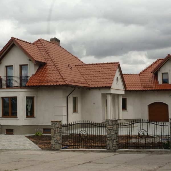 Dach domu pokryty dachówką ceramiczną Wienerberger Alegra 9 w kolorze czerwonej angoby. Budynek w jasnym kolorze, z prostą elewacją i ścieżką z kostki brukowej prowadzącą do wejścia.