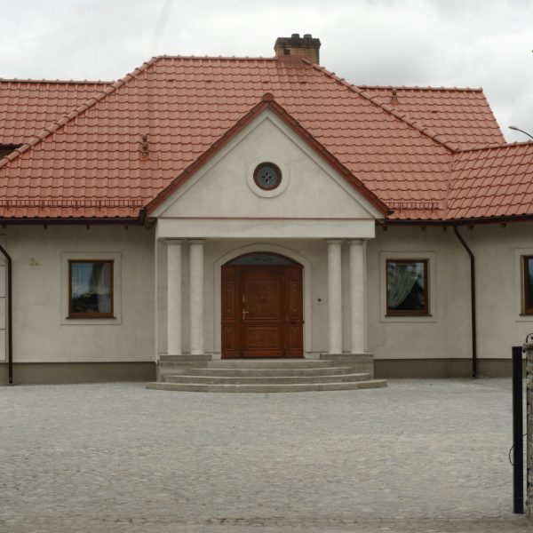 Dach domu pokryty dachówką ceramiczną Wienerberger Alegra 9 w kolorze czerwonej angoby. Budynek w jasnym kolorze, z prostą elewacją i ścieżką z kostki brukowej prowadzącą do wejścia.