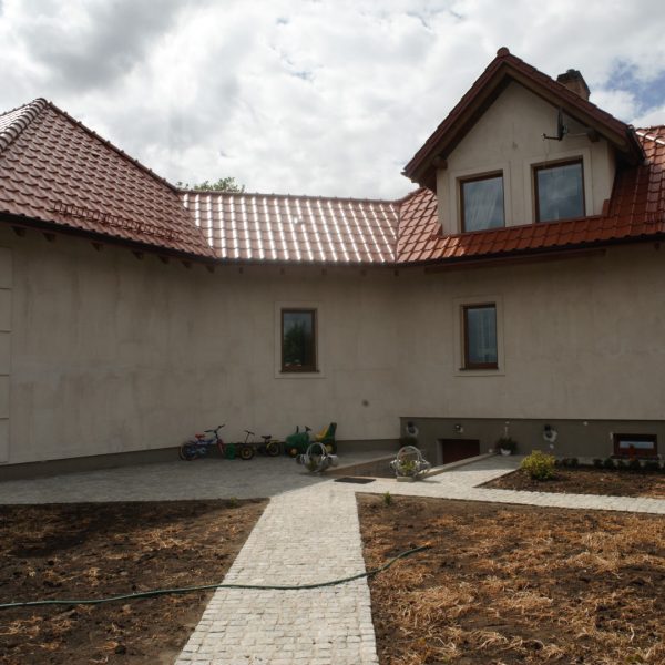 Dach domu pokryty dachówką ceramiczną Wienerberger Alegra 9 w kolorze czerwonej angoby. Budynek w jasnym kolorze, z prostą elewacją i ścieżką z kostki brukowej prowadzącą do wejścia.